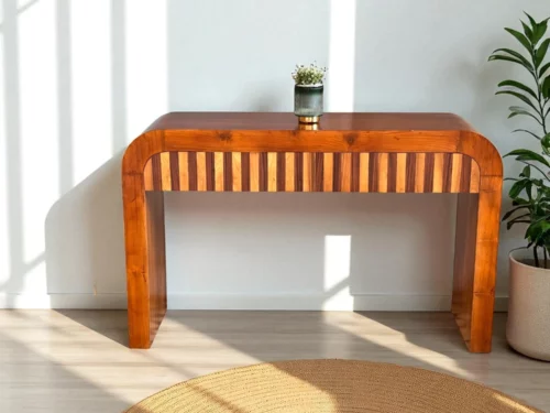 This elegant console table is crafted from premium Kerala Teak wood, featuring a sleek rectangular form with softly rounded edges and a natural matte finish that highlights the wood’s rich grain. The front panel showcases a striking vertical stripe pattern in alternating wood tones, adding visual depth and character. Thoughtfully integrated into the design are two concealed drawers, offering discreet storage without disrupting the clean aesthetic. Ideal for entryways, living rooms, or display corners, this piece blends craftsmanship, utility, and contemporary elegance—perfect for refined interiors.