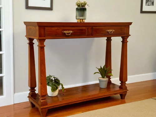 This Agara Console Table is made from very good, long-lasting teak wood from Nilambur (Kerala). It has a warm, deep brown color that shows off the natural wood grain. The table is very strong. It stands on four large, decorative wooden pillars for legs. It has two small drawers under the top for storing keys or small items, and they have nice, fancy brass handles. It’s an elegant, traditional piece that will look great in any home.