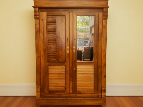 This elegant two-door wardrobe is crafted from premium Kerala Teak wood, showcasing a rich honey-brown finish that highlights the natural grain and warmth of the timber. The left door features a louvered panel with evenly spaced horizontal slats, adding texture and ventilation, while the lower section is framed with solid horizontal panels. The right door is fitted with a full-length mirror, perfect for daily use and enhancing room brightness, complemented by matching horizontal slats below. Fluted side columns and a crown molding at the top lend a regal touch, while brass-finished handles and keyholes add vintage charm. The wardrobe rests on a solid plinth base, offering stability and presence. Reflected in the mirror is a lively street scene, adding depth and character to the piece. Ideal for bedrooms or dressing areas, this wardrobe blends traditional craftsmanship with practical elegance—perfect for interiors that value heritage and utility.