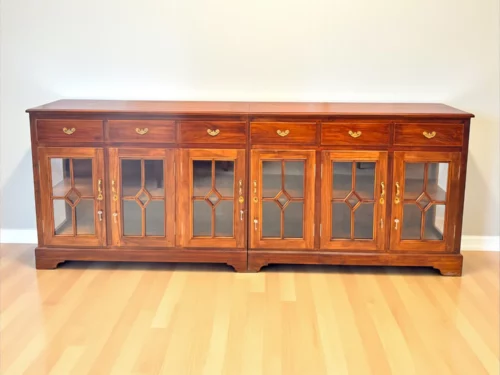 Large teak wood sideboard with six drawers and eight glass-paneled cabinet doors featuring geometric overlays; natural finish with brass handles, ideal for elegant dining or living room storage."