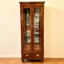 "Tall teak wood crockery shelf with glass doors, glass shelves, and brass shell handles on drawers; natural finish with rounded feet, ideal for elegant dining room display.